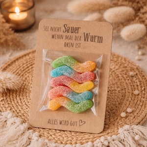Könnte beinhalten: Eine Karte mit deutschem Text, an der ein Päckchen bunter Gummibärchen befestigt ist. Die Gummibärchen befinden sich in einer durchsichtigen Plastiktüte. Auf der Karte steht der Satz "Alles wird gut" mit einem Herzsymbol. Die Würmer sind grün, blau, rot, orange und gelb.