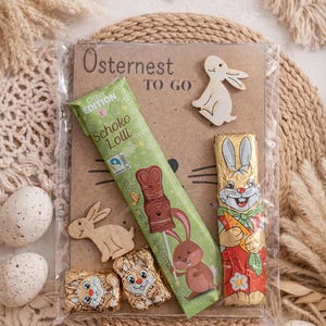 Puede incluir: Un set de regalo temático de Pascua con conejitos de chocolate, un Schoko Lolli y decoraciones de conejos de madera. El set se presenta sobre un salvamanteles tejido con huevos decorativos y hierba seca. El embalaje dice "Osternest TO GO."