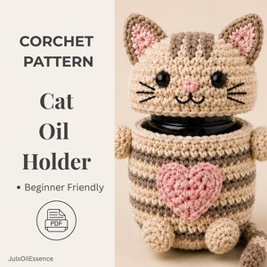 Könnte beinhalten: Häkelanleitung für einen katzenförmigen Ölhalter. Der handgefertigte Halter ist beige und braun mit einem rosa Herz und schwarzen Augen. Der Text auf dem Bild lautet "CORCHET PATTERN Cat Oil Holder" und "Beginner Friendly".