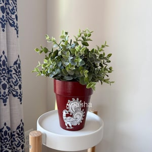 May include: A burgundy-colored flower pot with a white peacock design and the text "Apsha". The pot contains a green leafy plant. The pot is sitting on a white, round side table.