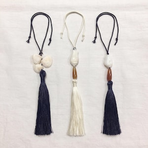May include: Three decorative hanging ornaments featuring tassels and shells. The left ornament has a dark navy tassel and three white fan shells. The centre has a white tassel, a white shell, and an amber bead. The right has a navy tassel, a white shell, and an amber bead.