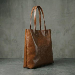Op de afbeelding: Een bruine leren tote bag met twee handvatten. De tas is rechthoekig met een glad, licht getextureerd oppervlak. De handvatten zijn dun en van hetzelfde leer. De tas is leeg en staat rechtop.