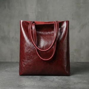 Op de afbeelding: Een bordeauxrode leren tote bag met een glanzende afwerking. De tas heeft twee handvatten en een rechthoekige vorm. De binnenkant is een lichtere tint rood. De tas staat op een grijze ondergrond.
