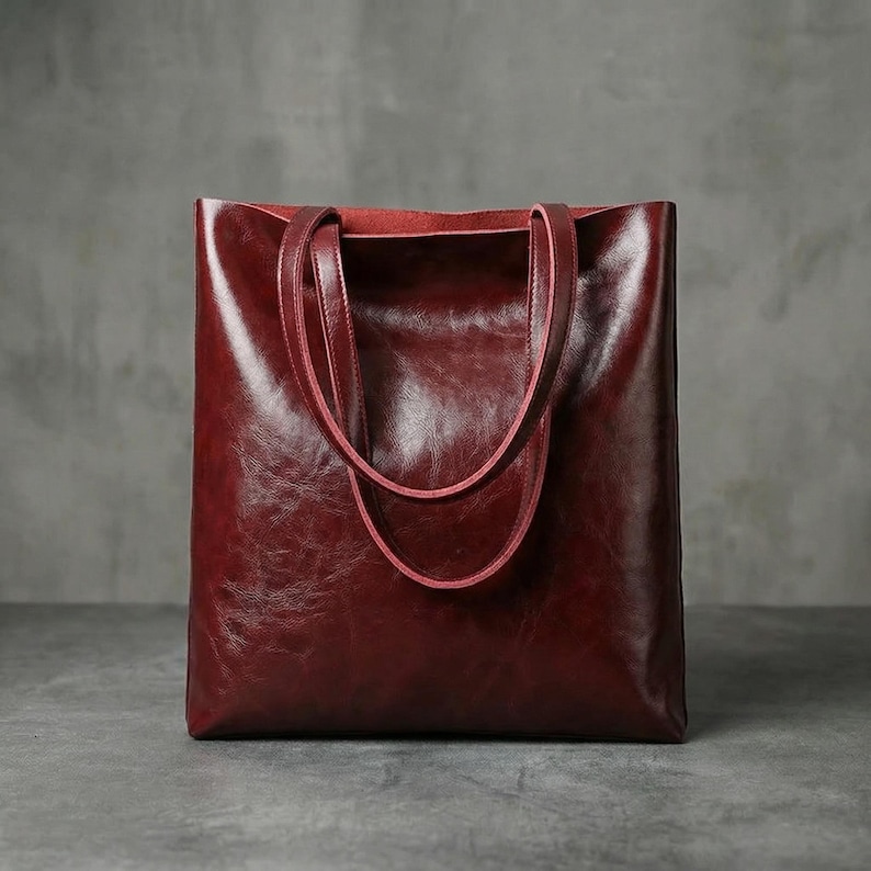 Op de afbeelding: Een bordeauxrode leren tote bag met een glanzende afwerking. De tas heeft twee handvatten en een rechthoekige vorm. De binnenkant is een lichtere tint rood. De tas staat op een grijze ondergrond.