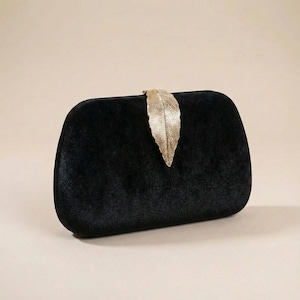 May include: Black velvet clutch with a gold-colored leaf clasp. The rectangular bag has rounded edges and a soft, textured appearance. The gold leaf clasp adds a touch of elegance to the design.