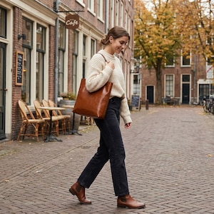 Op de afbeelding: Een persoon loopt over een geplaveide straat met een grote bruine leren draagtas. Ze draagt een cr&egrave;mekleurige trui, een zwarte jeans en bruine enkellaarsjes. De achtergrond toont gebouwen en een caf&eacute;.