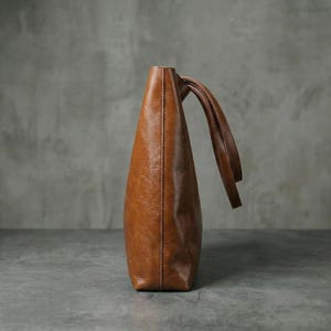Op de afbeelding: Een bruine leren tote bag met twee handvatten. De tas is een middelgroot formaat en heeft een eenvoudig, elegant ontwerp. Het leer heeft een licht verweerde uitstraling.