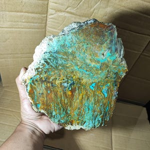 May include: A large, irregularly shaped stone with a striking pattern of teal, brown, and gold hues. The stone's surface has a textured appearance, with areas of white and black along the edges. The natural stone is held in a hand.