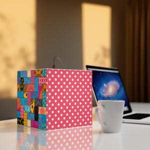 May include: A cube-shaped lamp with a patchwork design on one side and a pink polka dot pattern on another. The lamp is on a white surface, with a laptop and a white mug in the background.