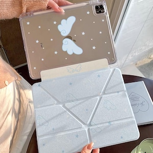 May include: Clear iPad case with a cloud and star design. The case is held open, revealing a light blue cover with a geometric pattern and cloud and star accents. A small book with text is also visible.