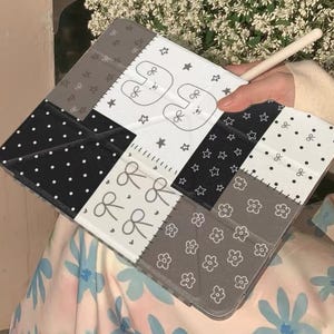 May include: A gray and white patchwork tablet case with a variety of patterns, including polka dots, stars, and floral designs. The case features a cute bunny illustration and the text "HXHStudio" at the bottom.