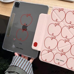 Op de afbeelding: Transparante iPad-hoes met een rood appelontwerp en de tekst "LINE APPLE". Een roze iPad-hoes met een herhalend rood appelomtrekpatroon is ook zichtbaar. De iPad-hoes wordt getoond op een houten oppervlak.