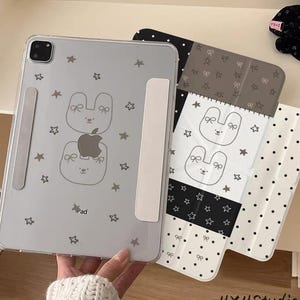 May include: Clear iPad case with a cartoon bunny design, stars, and the word "Pad". The case is shown with a black and white patterned cover. The bunny design includes a bow and an apple logo.