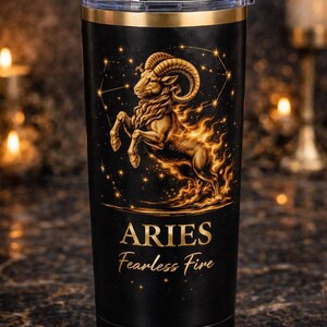 Puede incluir: Vaso negro con borde y tapa dorados. El diseño presenta un carnero dorado con llamas, constelaciones y el texto "ARIES Fearless Fire". El vaso es probablemente de acero inoxidable.