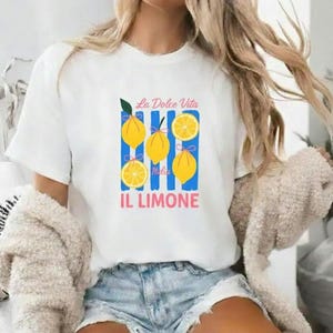 Può includere: T-shirt bianco con grafica colorata raffigurante limoni, strisce blu e la scritta italiana "La Dolce Vita Italia IL LIMONE". Il design include limoni interi e a fette con foglie verdi e fiocchi blu.