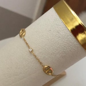 Peut inclure: Un bracelet doré délicat avec un design de chaîne. Le bracelet comporte deux maillons de forme ovale et une petite pierre précieuse étincelante. Le bracelet est présenté sur une surface blanche, avec une bande dorée en arrière-plan.