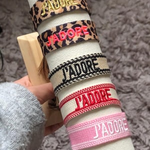 Può includere: Una collezione di braccialetti in tessuto con la scritta "J'ADORE" ricamata. I braccialetti presentano vari design, tra cui stampa leopardata e colori uniti come il rosa e il beige. I braccialetti sono esposti su un supporto cilindrico bianco.