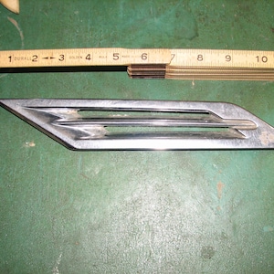 May include: A silver-colored, vintage car emblem with a streamlined design, featuring three horizontal bars and a pointed end. A wooden ruler, marked in inches, is placed above the emblem, indicating its size.
