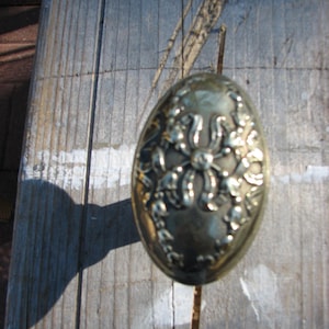 May include: Oval-shaped antique brass door knob with floral and ribbon detailing. The knob is set against a weathered wooden surface, casting a shadow. A decorative hardware element.