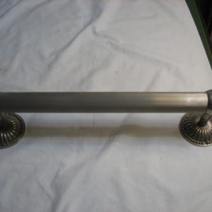 May include: A silver-toned metal towel bar with a ribbed center section and decorative, circular end mounts. The bar is approximately 12 inches long, with a polished, rounded end on each side. The end mounts have a detailed, scalloped design.