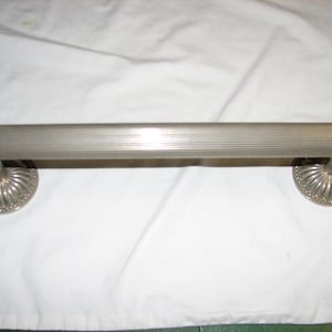 May include: A silver-toned towel bar with a ribbed center section and rounded end caps. The bar is supported by decorative, circular bases with a scalloped edge. The item appears to be made of metal and is designed for bathroom use.