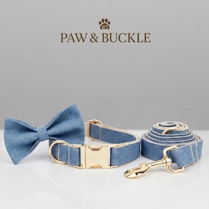 May include: A denim dog collar, leash, and bow tie set with gold-colored hardware. The bow tie is tied, and the leash is coiled. The set is branded with the text "PAW & BUCKLE" in brown.