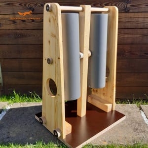 May include: A wooden stand featuring two vertical gray cylindrical rollers suspended between three light-colored wooden uprights. The structure is mounted on a dark brown rectangular base, with circular cutouts in the side panels and small circular accents.