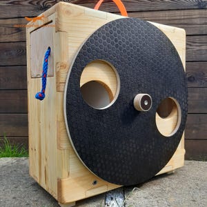 May include: A light-colored wooden box with a large, circular black disc featuring a textured honeycomb pattern and two circular cutouts. A small wooden knob is centered on the disc. A blue braided rope hangs from the side, and an orange handle is on top.