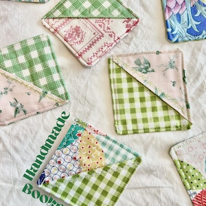 May include: Handmade fabric bookmarks in various patterns, including green gingham, floral, and cross-stitch designs. The bookmarks are square with a diagonal fold, and the text "Handmade Bookmarks" is printed on the fabric.