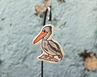 May include: A ceramic pelican ornament featuring a cream-coloured body with grey shading, a bright orange beak, and orange feet. The bird is depicted standing on a small grey base and is suspended from a thin, dark metal hook for display purposes.
