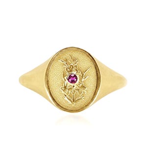 May include: A gold signet ring with an oval face. The face features a floral design with a small, round, red gemstone at the center. The ring has a smooth, polished finish and a classic, elegant design.