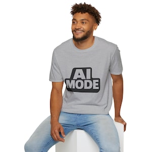 May include: A light gray t-shirt with the text "AI MODE" in a black, rectangular design. The shirt is short-sleeved and appears to be made of a soft material. The person in the image is wearing blue jeans.