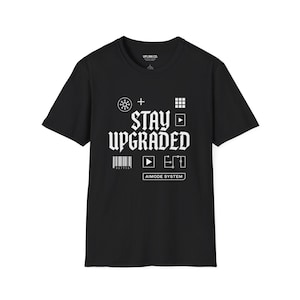 May include: Black t-shirt featuring the text "STAY UPGRADED" in a bold, stylized font, accompanied by white graphic symbols. The words "AIMODE SYSTEM" are printed below. The shirt has a crew neck and short sleeves.
