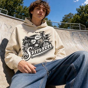 Op de afbeelding: Een beige hoodie met een zwart-witte grafische illustratie van een Simson-motorfiets op de voorzijde. Het ontwerp bevat het woord Simson in een gestileerd scriptlettertype onder de motorfiets, met een gestippeld halftoon-achtergrondeffect.