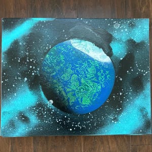 May include: A canvas painting depicting a planet in space. The planet is primarily blue and green, with a white area on the upper right. The background is black with turquoise spray paint and white dots, suggesting stars and nebulae. The artwork is a vibrant representation of a celestial body.