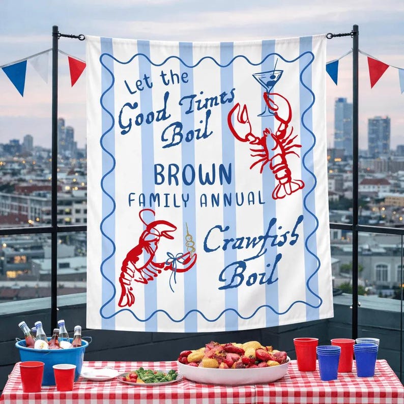 May include: A white banner with blue stripes and text that reads "Let the Good Times Boil BROWN FAMILY ANNUAL Crawfish Boil." Red crawfish and a martini glass are illustrated. The banner is hanging above a table set for a crawfish boil.
