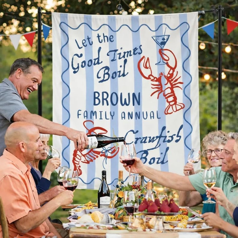 May include: A festive outdoor gathering with a banner that reads "Let the Good Times Boil BROWN FAMILY ANNUAL Crawfish Boil." People are gathered around a table, enjoying wine and food. The banner features a lobster and a cocktail.