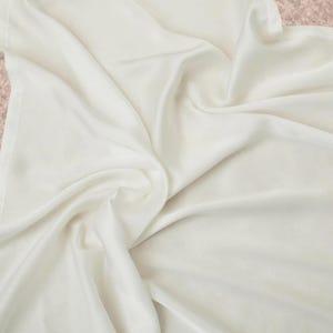 May include: Off-white fabric with soft folds and a silky texture. The fabric is draped, creating shadows and highlights that emphasize its smooth surface. The overall appearance suggests a luxurious textile.