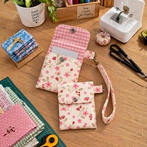 May include: A cream-colored fabric wallet and a matching smaller pouch, both featuring a pink and red floral print. The wallet has a pink gingham interior lining and a wrist strap. Both items are secured with silver-toned metal snap buttons.