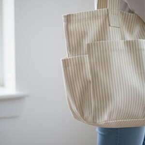 May include: A beige and white striped tote bag with an external pocket. The bag is made of a sturdy fabric and has a simple design. The tote bag is held by a person.