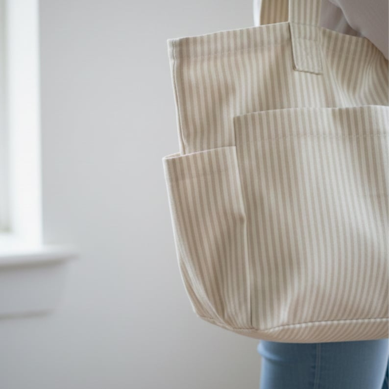 May include: A beige and white striped tote bag with an external pocket. The bag is made of a sturdy fabric and has a simple design. The tote bag is held by a person.