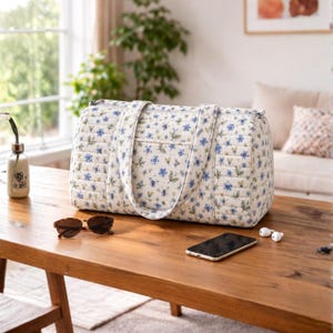 May include: A quilted duffel bag featuring a white base with a repeating pattern of small blue flowers and green leaves. The bag has two long shoulder straps, a rectangular shape, and a zippered top closure with a soft, padded texture.
