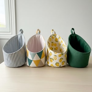May include: Four fabric hanging storage baskets. The baskets feature various patterns: blue and white stripes, geometric shapes in yellow, green, and grey, lemons on a yellow background, and solid green. Each has a loop for hanging.
