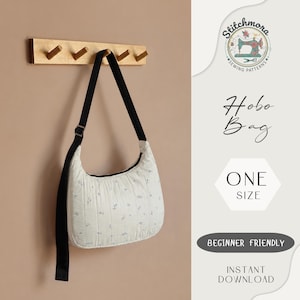 Pode incluir: Uma bolsa hobo branca com um pequeno padrão floral azul e uma alça preta ajustável pendurada num gancho de parede de madeira. O texto Stitchmora Sewing Patterns, Hobo Bag, One Size, Beginner Friendly e Instant Download é visível.
