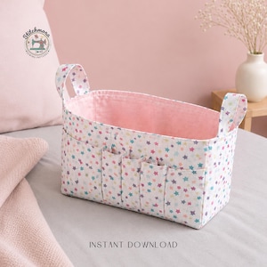 Peut inclure: Un panier de rangement en tissu avec un extérieur blanc orné d'étoiles colorées. L'intérieur est rose et comporte plusieurs petites poches à l'extérieur. Le panier a deux poignées et le texte "INSTANT DOWNLOAD" est imprimé en bas.