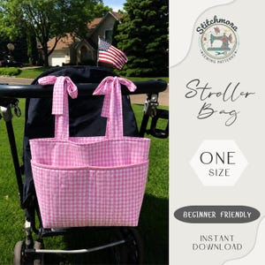 Puede incluir: Un bolso para cochecito de bebé con estampado de cuadros vichy rosa y blanco, con dos asas atadas en lazos y un bolsillo frontal acolchado. El bolso está sujeto a un manillar negro. El texto a la derecha dice Stitchmora Stroller Bag ONE SIZE.