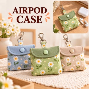 May include: Three square fabric Airpod cases with daisy patterns in blue, green, and beige. Each case features a flap with a snap button closure, a metal lobster clasp, and a small daisy print with tiny hearts on a soft textile surface.