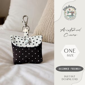 May include: A small fabric pouch with a black and white polka dot pattern, featuring a fold-over flap secured by a white snap button. A silver metal lobster clasp is attached to the top via a fabric loop. The text reads AirPod Cover ONE SIZE.