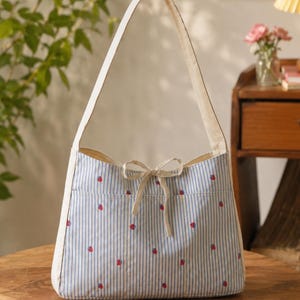 Puede incluir: Un bolso de hombro con un patrón de rayas verticales en azul claro y blanco con pequeños motivos de fresas rojas bordadas. El bolso incluye una correa de color blanco roto, un cierre de lazo a juego en el centro y el texto STEP BY STEP GUIDE BEGINNER FRIENDLY.