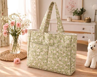 May include: A sage green tote bag featuring a repeating pattern of small white and pink flowers. The bag has two top handles and two exterior front pockets. The fabric has a structured rectangular shape with visible stitching along the seams.